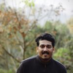 A smiling young South Asian man in a black jacket enjoying the outdoors in fall.